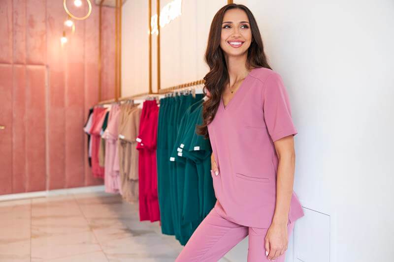 Kobieta w różowym uniformie medycznym na tle wieszaków z odzieżą medyczną.
