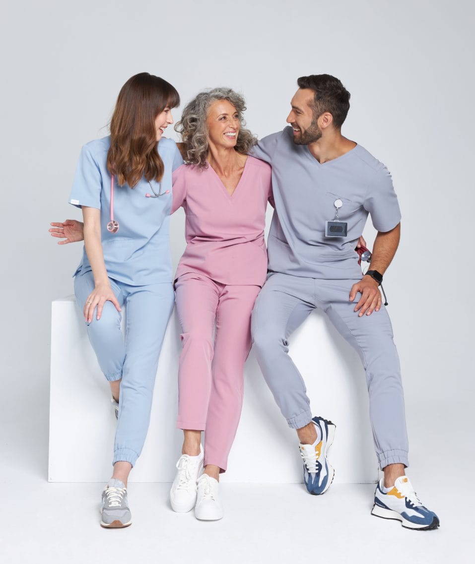 Trzy uśmiechnięte osoby w kolorowych ubraniach medycznych siedzące razem i rozmawiające.