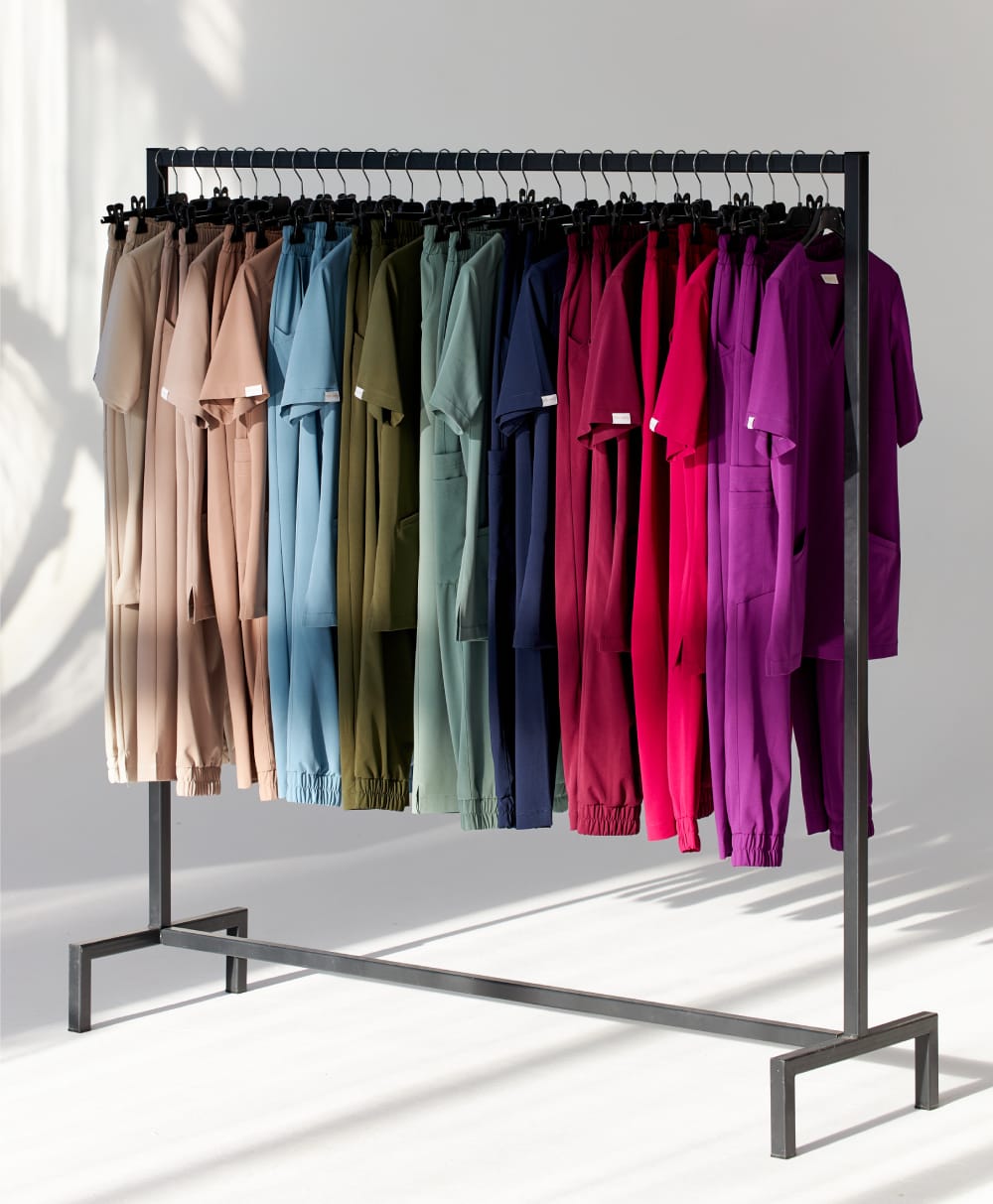 Kolorowe zestawy odzieży medycznej na wieszaku w różnych odcieniach beżu, niebieskiego, zielonego, granatu i fuksji.