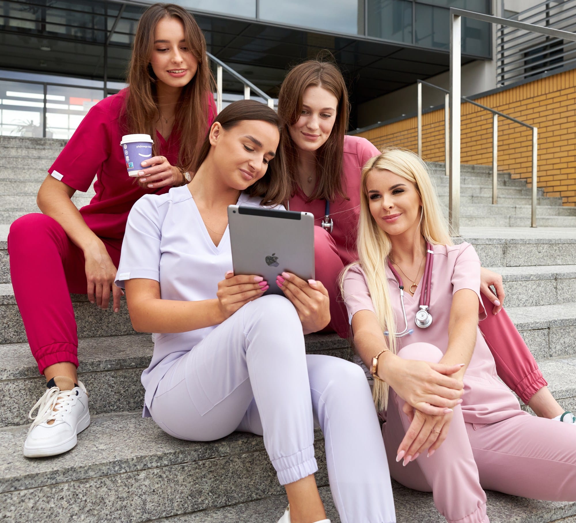 Cztery uśmiechnięte kobiety w kolorowej odzieży medycznej siedzą na schodach i korzystają z tabletu.