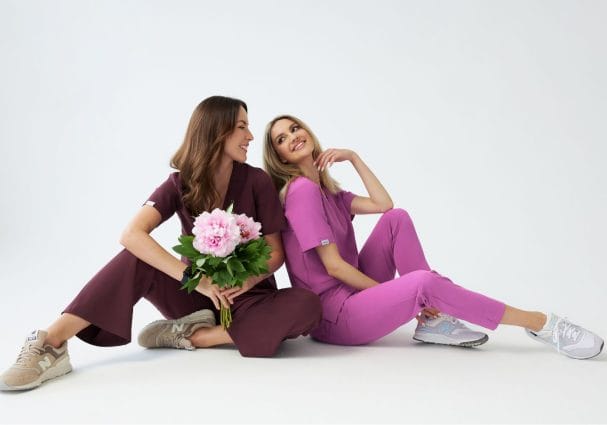 Dwie uśmiechnięte kobiety w kolorowej odzieży medycznej siedzą na podłodze, jedna trzyma bukiet kwiatów.