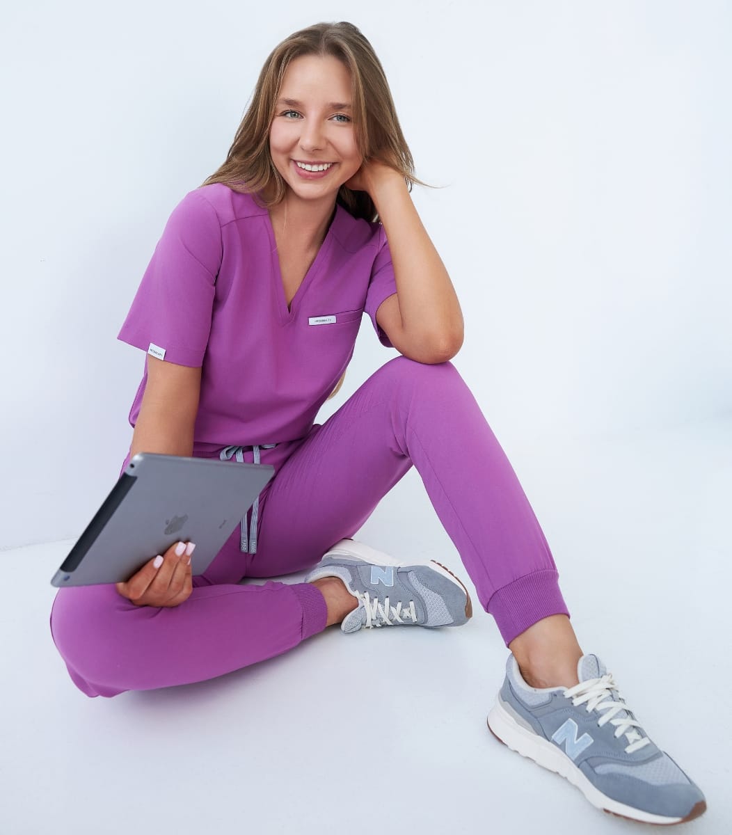 Młoda kobieta w fioletowej odzieży medycznej siedzi na podłodze z tabletem i uśmiecha się.