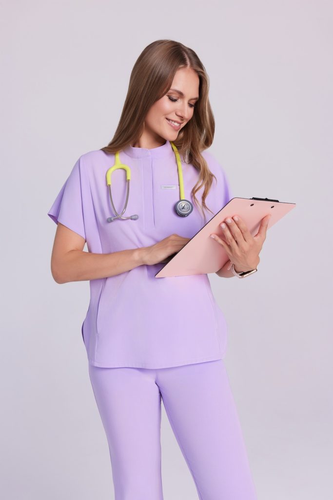 Kobieta w liliowej bluzce medycznej scrubs Basic Flex trzymająca clipboard i stetoskop